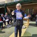 Starejša gospa na prostem drži priznanje in medaljo za tretje mesto, v ozadju sedijo drugi stanovalci in spremljajo dogajanje.