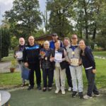 Skupina osmih ljudi na prostem ponosno pozira s priznanji in medaljami po športnih igrah, v ozadju so drevesa in lesena uta.