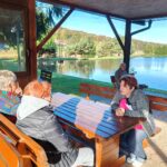 Skupina žensk in en moški sedijo pod nadstreškom ob ribniku in se sproščeno pogovarjajo.