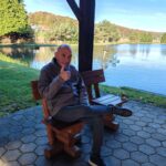 Moški sedi na klopi ob ribniku in z dvignjenim palcem kaže dobro voljo.