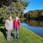 Dva moška stojita ob vodi, sončen dan pa osvetljuje jesensko pokrajino.