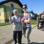 Starejša gospa in mlajša spremljevalka hodita nasmejani ob cesti. Gospa se naslanja na roko spremljevalke, za njima pa se vidijo drugi udeleženci sprehoda.