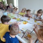 Otroci in stanovalci skupaj ustvarjajo z barvami pri skupni mizi, na listih nastajajo pisani odtisi.