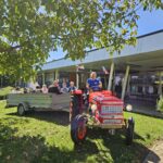 Rdeč traktor z vozom, v katerem sedijo ljudje, stoji pred domom. Voznik se smehlja, na vozilu pa so pripete slovenske zastave.