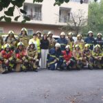 Skupina gasilcev v popolni zaščitni opremi in s čeladami stoji in kleči pred domom za starejše. Dve zaposleni stojita med njimi. Fotografija je posneta po gasilski vaji evakuacije.
