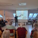 Predavatelj stoji pred projekcijskim platnom in razlaga postopke ob nevarnosti požara stanovalcem doma za starejše, ki sedijo v dvorani.