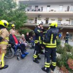 Gasilci pomagajo evakuiranim stanovalcem doma za starejše, ki sedijo zunaj na invalidskih vozičkih. V ozadju se vidi stavba doma in drugi stanovalci na balkonih.
