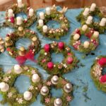 Na modri podlagi je razporejenih več adventnih venčkov iz mahu, okrašenih z rdečimi, belimi, zlatimi in srebrnimi svečami ter okrasnimi kroglicami in naravnimi dodatki.