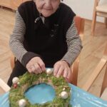 Starejša ženska sedi za mizo in okrašuje adventni venček iz mahu z velikimi svetlečimi okrasnimi kroglicami; pred njo je zaščitna modra podlaga.
