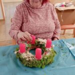 Ženska z rdečimi lasmi okrašuje adventni venček, na katerega namešča živo rožnate sveče in biserne okraske; okoli nje so materiali za ustvarjanje.