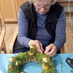 Starejša ženska s svetlimi lasmi pritrjuje rdeče okraske na adventni venček, ki ima štiri zlate sveče; pred njo je modra zaščitna podlaga in ustvarjalni material.