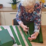 Gospa reže zelene trakove iz krep papirja za izdelavo cvetličnega aranžmaja. Na mizi so tudi košara, goba za cvetje in ostanki materiala.