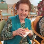 Stanovalka z nasmehom drži sendvič in uživa ob prijetnem druženju.