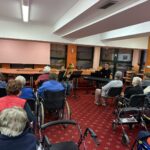 Stanovalci sedijo v dvorani in poslušajo mladega glasbenika, ki igra na trobento ob spremljavi klaviature.