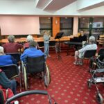 Fant stoji pred občinstvom in igra trobento, ob strani pa ga spremlja pianist; stanovalci pozorno spremljajo nastop.