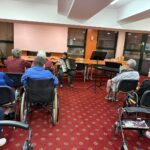 Dekle sedi pred občinstvom in igra harmoniko, stanovalci pa z zanimanjem opazujejo glasbeni nastop.