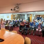 Polna dvorana stanovalcev pozorno spremlja mlado glasbenico, ki ob klavirski spremljavi igra trobento.