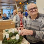 Starejši gospod z nasmehom drži adventni venček iz zelenja ter nanj namešča bele sveče; ob njem sedijo drugi stanovalci, ki prav tako ustvarjajo.