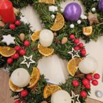 Več adventnih venčkov iz zelenih vejic, okrašenih z belimi in vijoličnimi svečami, suhimi pomarančnimi rezinami, zvezdicami in rdečimi jagodami, razporejenih na mizi.