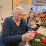 Starejša gospa sedi za mizo in na adventni venček iz zelenja namešča rdečo svečo, medtem ko druga stanovalka ob njej z nasmehom opazuje.