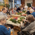 Skupina starejših oseb sedi okoli dolge mize in okrašuje adventne venčke z zelenjem, svečami in okrasnimi materiali v skupnem prostoru doma.