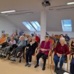 Skupina stanovalcev sedi in spremlja dogodek v skupnem prostoru doma.