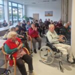 Stanovalci doma sede in v invalidskih vozičkih spremljajo nastop zunanje skupine ter navdušeno ploskajo.