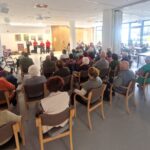 Večja skupina stanovalcev v dvorani doma spremlja nastop zunanje glasbene skupine na odru.