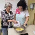 Stanovalka s pomočjo zaposlene meša maso za peko v veliki posodi.