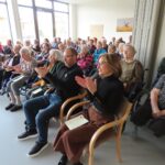 Občinstvo, ki ga večinoma sestavljajo stanovalci doma, sedi in navdušeno ploska med nastopom.