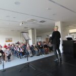 Pevka ob spremljavi pianista nastopa na odru pred polno dvorano stanovalcev.