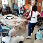 Stanovalci sedijo okoli mize in poslušajo igranje harmonike med peko pogače.