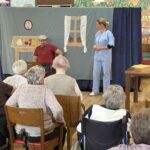 Pred obiskovalci doma poteka humoristična gledališka predstava z igralcem in igralko v scenski postavitvi kuhinje.