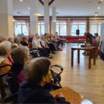 Stanovalci doma spremljajo gledališko predstavo, ki poteka pred njimi v jedilnici.