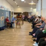 Igralka v modri opravi pomaga igralcu na odru, medtem ko občinstvo pozorno spremlja prizor.