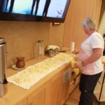 Zaposlena pripravlja narezane krompirjeve palčke, razporejene na kuhinjskem pultu, ob njej pa so kuhinjski pripomočki in posode za kuhanje.