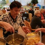 Starejša gospa z zajemalko pobira ocvrt krompirček iz posode z vročim oljem, medtem ko druga oseba drži posodo za serviranje; v ozadju sedijo stanovalci.