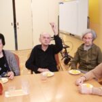 Štirje stanovalci sedijo za mizo, pred seboj imajo krožničke s krompirčkom; eden izmed njih dvigne kos krompirčka v roki.