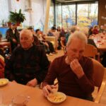 Skupina starejših oseb sedi za večjo jedilno mizo in je krompirček, nekateri opazujejo dogajanje, drugi se pogovarjajo v prijetnem skupnem prostoru doma.