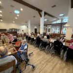 Stanovalci sedijo za pogrnjenimi mizami in skupaj praznujejo rojstni dan v jedilnici doma.