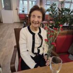 Stanovalka z nasmehom sedi za mizo in drži šopek rdečih nageljnov.