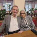 Stanovalec in stanovalka sedita drug ob drugem ter se nasmihata pred praznično okrašenim prostorom.