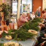 Skupina starejših oseb sedi ob dolgi mizi in oblikuje adventne venčke iz zelenja in slame v skupnem prostoru doma.