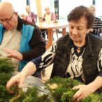 Starejša gospa in moški skupaj oblikujeta adventne venčke iz zelenih vejic, medtem ko drugi stanovalci spremljajo dogajanje.