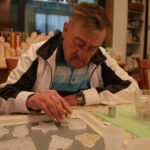 Starejši gospod posipa bleščice po prazničnih okraskih iz silikonskih modelčkov na mizi.