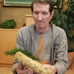 Starejši moški drži slamnato osnovo adventnega venčka in nanjo pritrjuje zelene vejice.