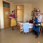 Obisk klovnov v sobi, kjer z glasbo in humorjem razveseljujeta stanovalca na postelji.