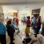 Skupina stanovalcev se v hodniku zbere okoli klovnov in spremlja njun nastop.