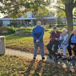 Skupina starejših oseb sedi na klopeh v jesensko obarvanem parku doma za starejše; nekateri uporabljajo hodulje, drugi klepetajo, moški stoji na poti med njima, sonce pa sveti skozi drevesa v ozadju.