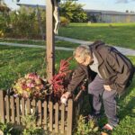 Starejši moški v parku pri križu s strešico polaga svečo v lesen cvetlični okvir, ki je okrašen z jesenskim cvetjem; prizor je obsijan s toplim popoldanskim soncem.